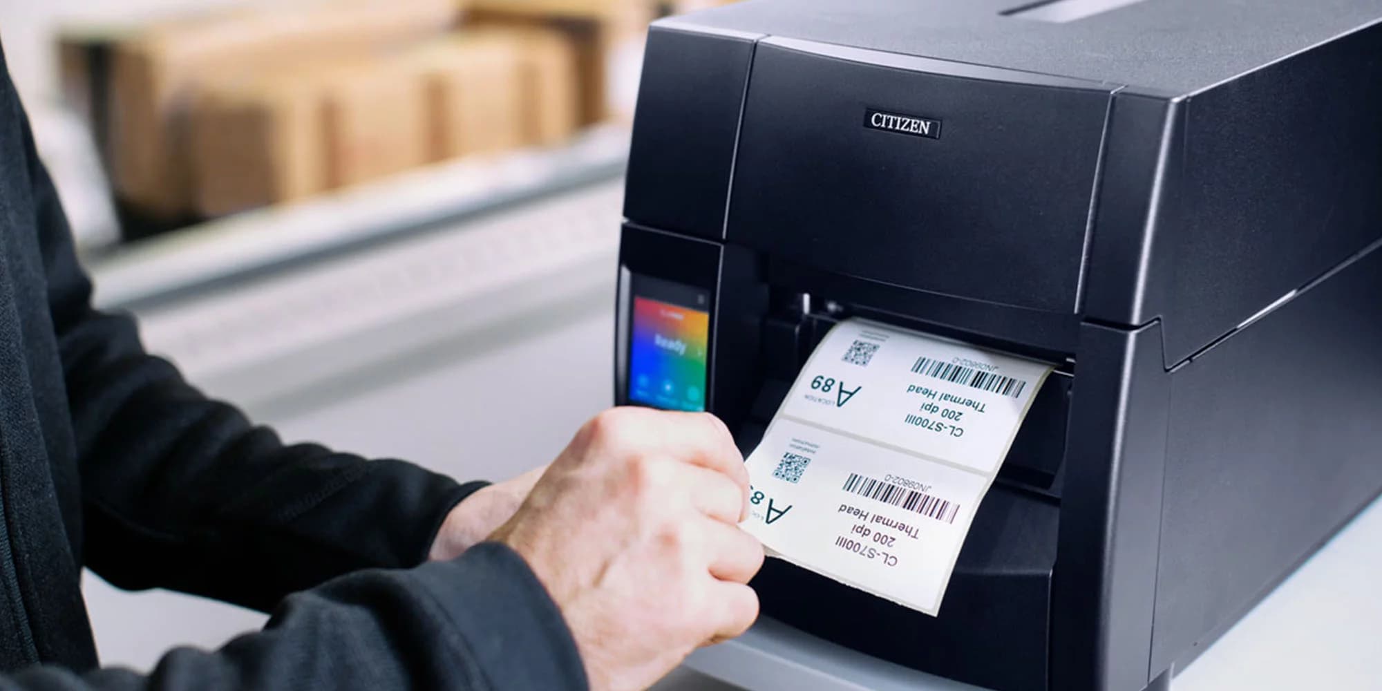 Hands removing a printed barcode label from a Citizen industrial printer in a warehouse setting.