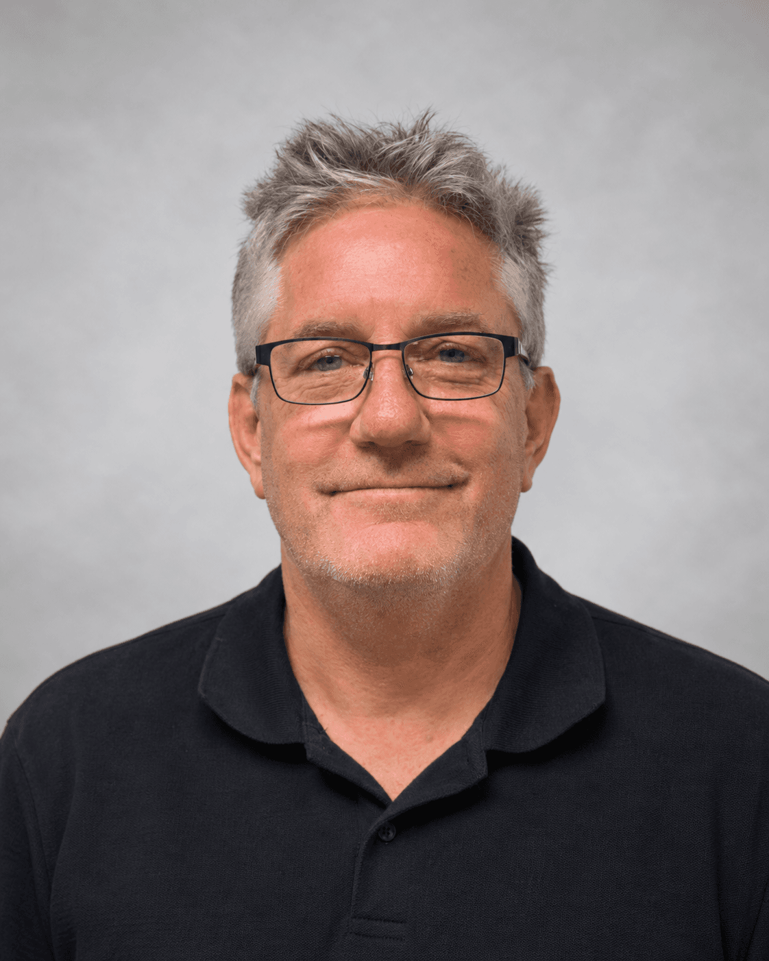 Smiling middle-aged man with messy grey hair and glasses wearing a black polo shirt.