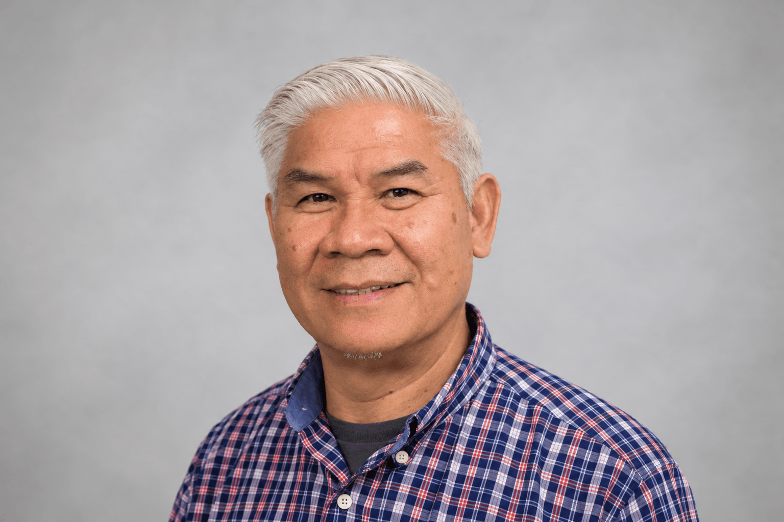 Smiling older Asian man with short white hair wearing a blue and red plaid shirt.