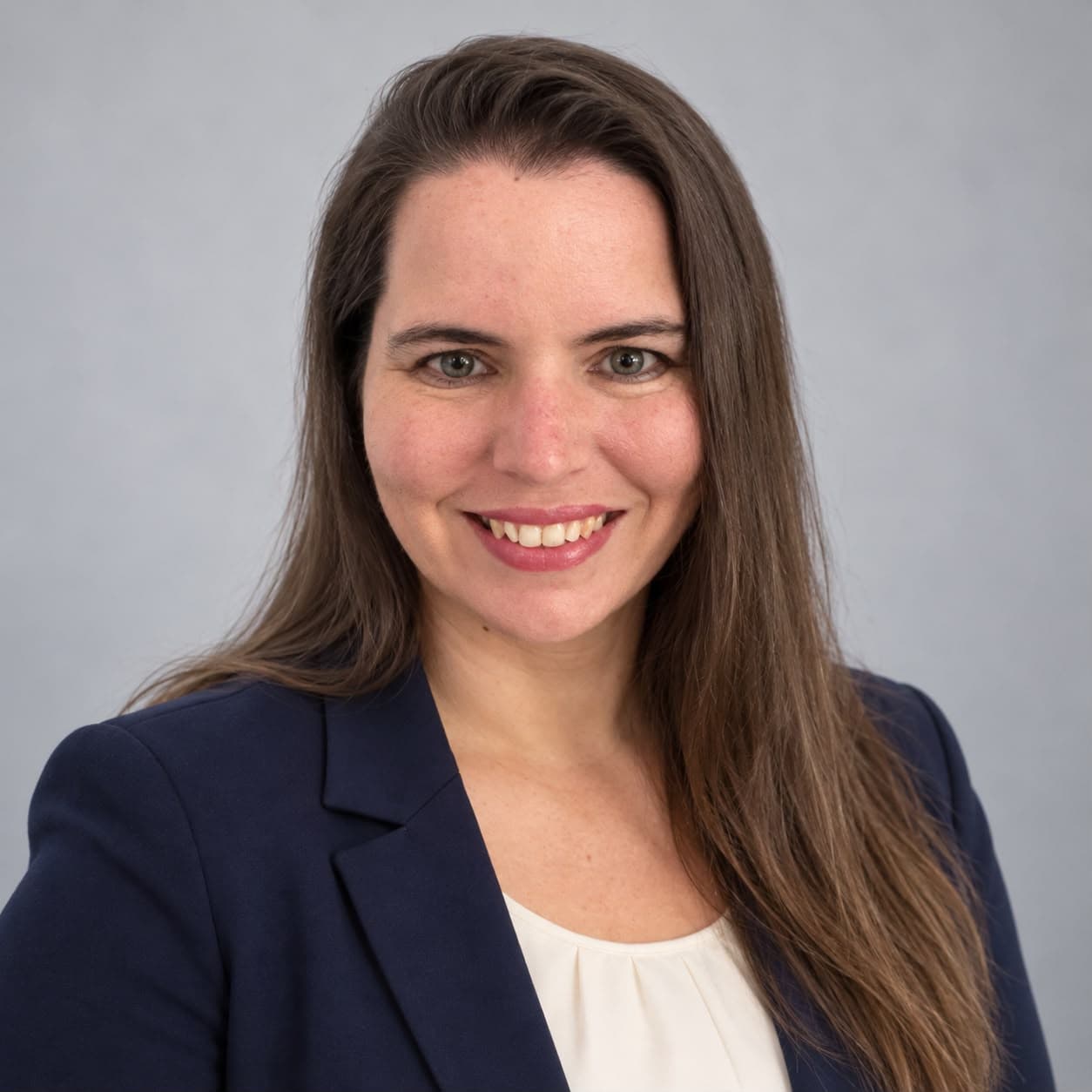 Smiling woman with long brown hair wearing a navy blue blazer and white blouse.