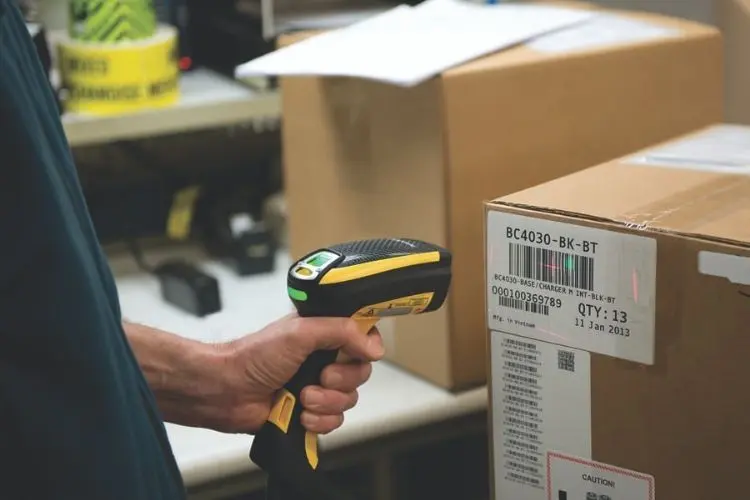 Handheld barcode scanner scanning a shipping label on a cardboard box in a warehouse.