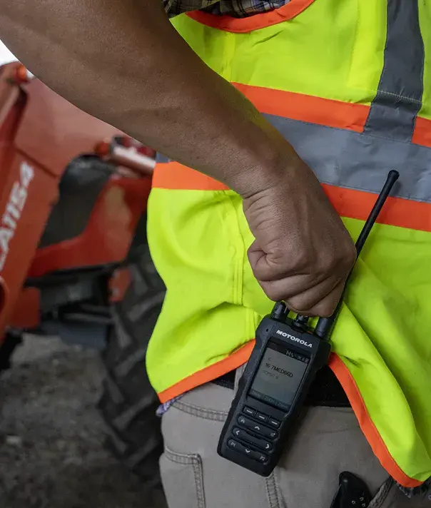 Worker in a high-visibility safety vest holds a Motorola radio near heavy construction equipment.