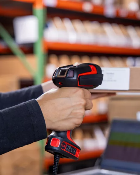 Handheld barcode scanner scanning a white label on a cardboard box in a warehouse.