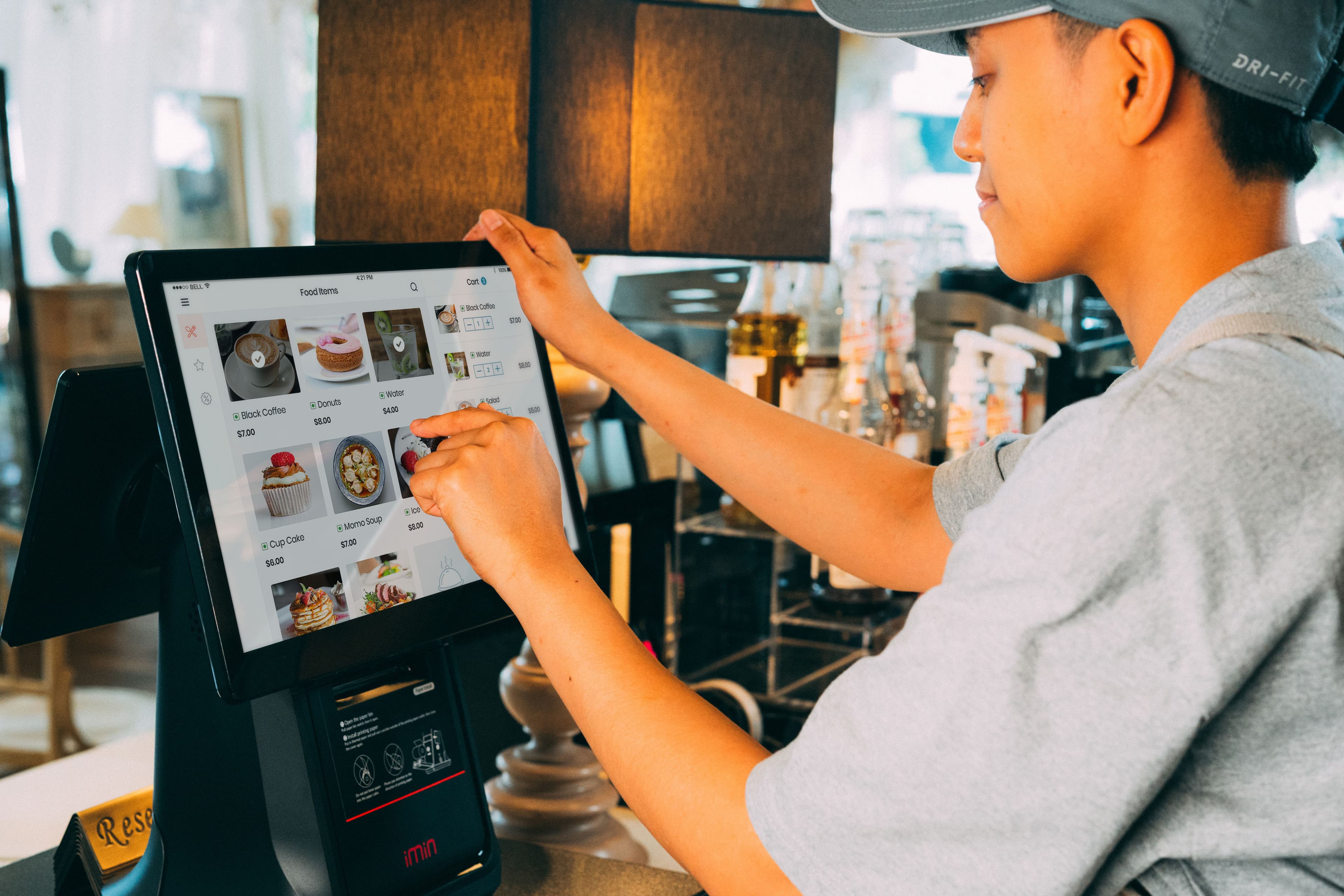 A cafe worker selects food items on a touch-screen point-of-sale system at a counter.