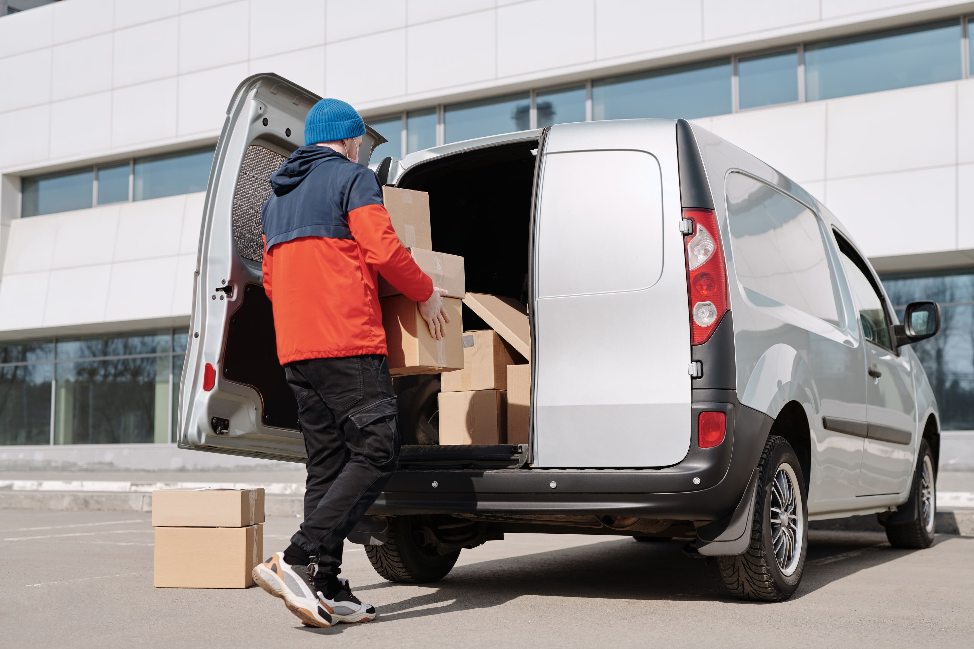 A courier loading cardboard boxes into the open back of a silver delivery van.