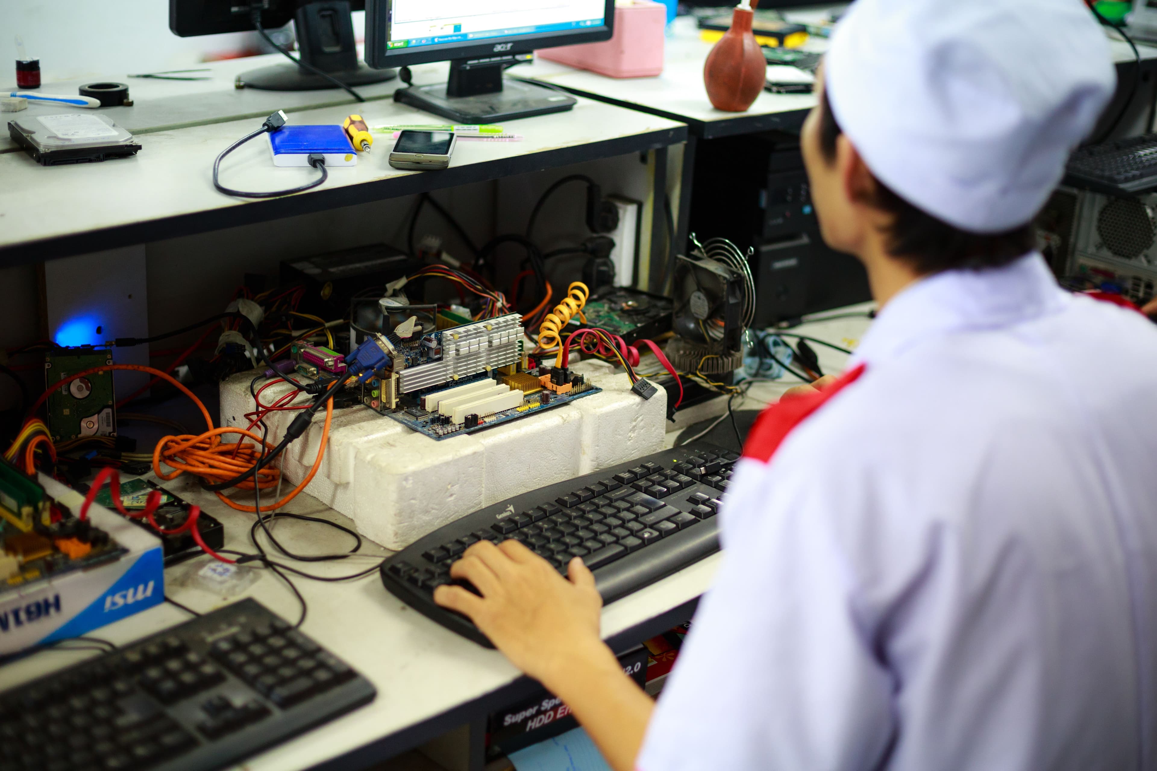 Technician in a white cap repairs an exposed motherboard at a cluttered electronics repair workstation.