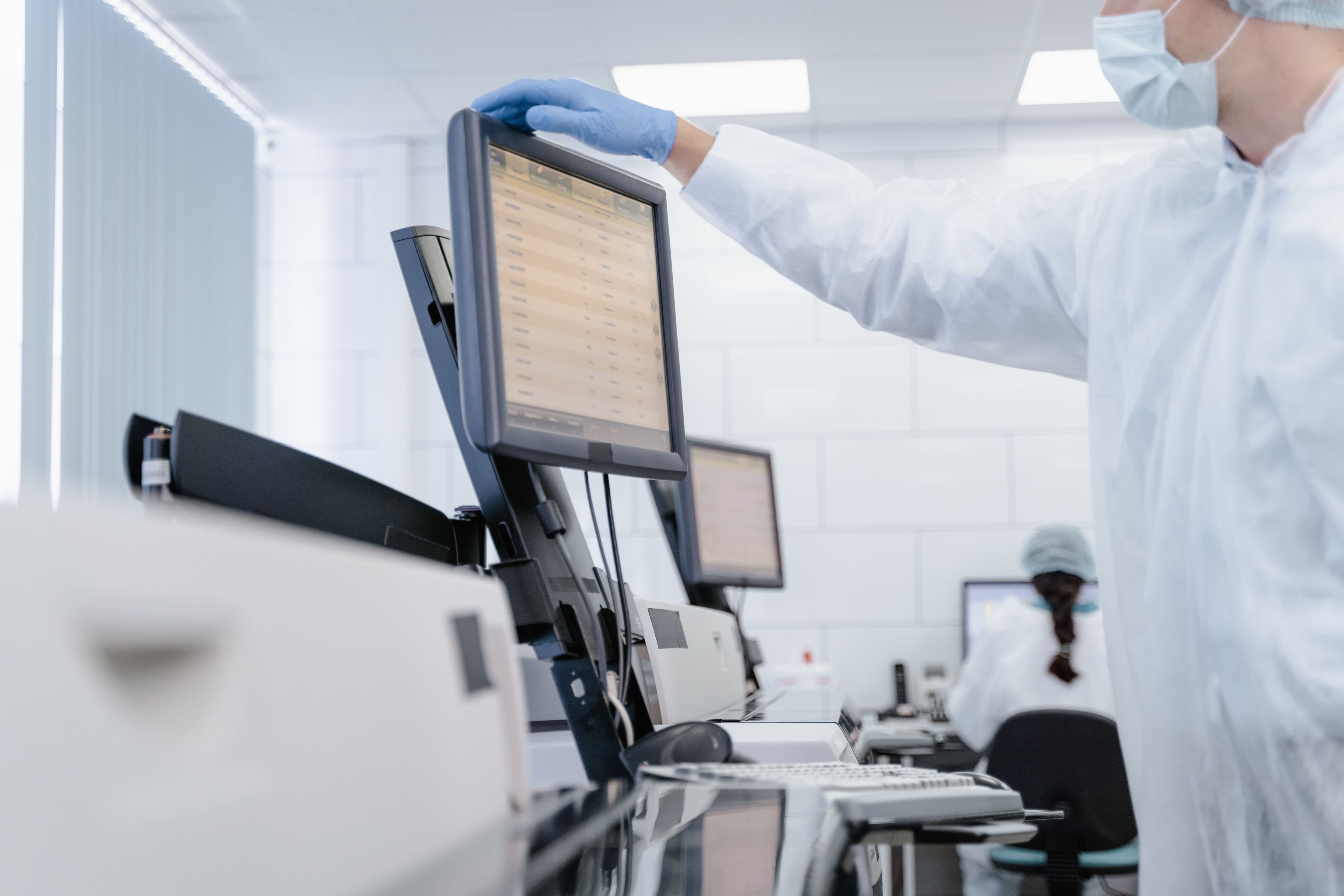 Technician in protective gear adjusts a computer monitor in a clean, sterile medical laboratory.